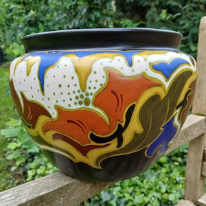 Könnte beinhalten: Ein großer, runder Keramiktopf mit schwarzem Hintergrund und einem farbenfrohen Blumendesign. Das Design zeigt orange, rote, gelbe, blaue und weiße Blumen und Blätter.