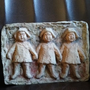 Puede incluir: Una escultura en relieve de terracota de tres niños de pie uno al lado del otro. Los niños visten ropa sencilla y tienen caras redondas. La escultura es de color marrón claro y tiene una textura rugosa.