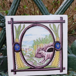 May include: A vintage ceramic tile with a hand-painted design. The tile features a white wolf in a forest setting, surrounded by green foliage and a blue sky. The wolf is looking to the right, and there are three smaller animals in the background. The tile is framed by a purple and black border with yellow and green accents.