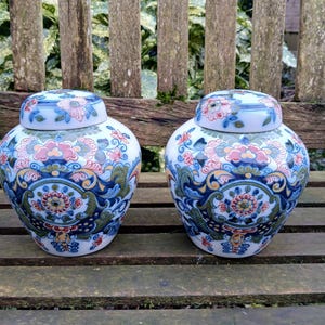 Könnte beinhalten: Zwei weiße Keramik-Behälter mit blauen, grünen und roten floralen Mustern. Die Behälter haben Deckel und stehen auf einer Holzoberfläche.