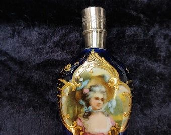 Może przedstawiać: Szklana butelka perfumowa w kolorze kobaltowym z srebrnym korkiem i złotymi akcentami. Butelka zdobiona jest ręcznie malowanym portretem kobiety w białym kapeluszu i różowej sukience.