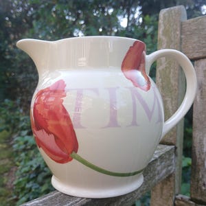 May include: Cream ceramic jug featuring a red tulip design and the word "TIME" in lavender. The jug has a curved handle and a pouring spout. The floral design adds a touch of spring to any kitchen or dining room.