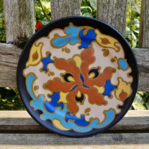 Op de afbeelding: Een decoratieve keramische plaat met een zwarte rand en een centraal bloemmotief. Het ontwerp heeft een crèmekleurige achtergrond met oranje, blauwe en gele bloempatronen. De plaat is rond en lijkt vintage.