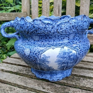 Op de afbeelding: Een grote blauwe en witte keramische plantenpot met twee handvatten. De pot heeft een decoratief ontwerp met een landschapsscène met bomen en een geometrisch patroon.
