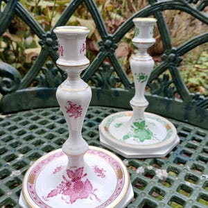 May include: Two white porcelain candlesticks with floral designs in pink and green. The candlesticks have gold trim and are set on a hexagonal base. The pink candlestick has a detailed floral pattern, while the green candlestick has a simpler design.