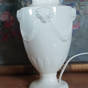 May include: A white ceramic table lamp with a classic urn shape. The lamp features decorative ram's head accents and a fluted base. The lamp is approximately 15 inches tall and has a white cord.