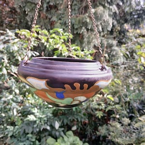 May include: A hanging ceramic planter with a dark brown exterior and decorative painted design in orange, blue, and green. The planter is suspended by a chain, with a backdrop of green foliage.