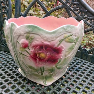 Puede incluir: Una maceta decorativa de cerámica con borde festoneado y diseño floral. El interior es rosa claro, el exterior crema con una gran flor burdeos y hojas verdes. La maceta está sobre un banco de metal verde.