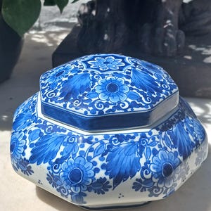 Peut inclure: Un pot en céramique hexagonal avec couvercle, présentant une base blanche ornée de motifs floraux et de feuilles bleus complexes. Le design recouvre à la fois le couvercle et le corps du pot, affichant une disposition symétrique de fleurs.