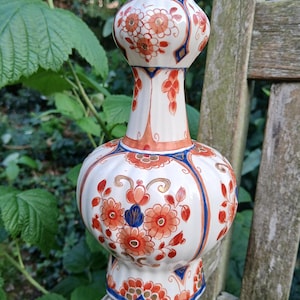 May include: A white ceramic vase adorned with orange and blue floral patterns. The vase features a rounded body, a narrow neck, and a flared rim. The floral design incorporates orange flowers, blue accents, and gold detailing. It is a decorative item.