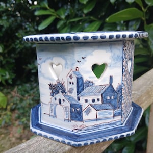 May include: A blue and white ceramic candle holder with a hexagonal shape and cut-out hearts. The holder features a painted scene of a village with houses and trees.