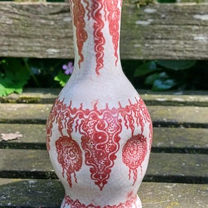 Könnte beinhalten: Eine weiße Keramikvase mit roten dekorativen Mustern. Die Vase hat einen bauchigen Körper, einen schlanken Hals und einen ausgestellten Rand. Die roten Muster umfassen florale und abstrakte Designs und verleihen dem Stück einen Hauch von Eleganz.