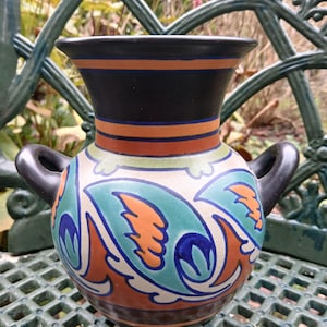 Könnte beinhalten: Eine Keramikvase mit schwarzem Rand und Griffen. Die Vase hat einen cremefarbenen Körper mit teal-, orange- und blaufarbenen Mustern. Die Vase steht auf einer grünen Metalloberfläche. Höhe ca. 15 cm.