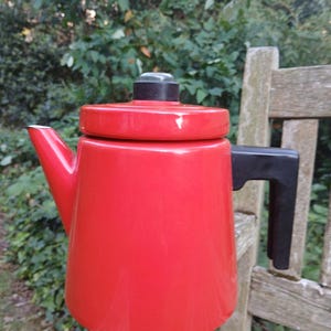 Puede incluir: Una tetera roja vibrante con asa, tapa y boquilla negras. La tetera es cilíndrica con base redondeada y está sobre una superficie de madera. El fondo presenta un exuberante follaje verde.