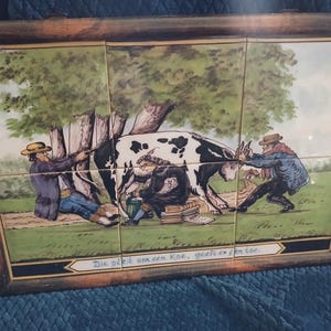 May include: A framed tile artwork depicting a pastoral scene with a black and white cow, three figures, and a tree. The artwork is composed of multiple tiles and features a wooden frame. The scene includes text at the bottom.