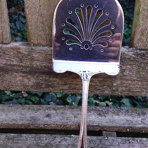 Puede incluir: Un utensilio de servir de plata con un diseño decorativo de concha. El utensilio tiene un mango largo con un borde de cuentas.