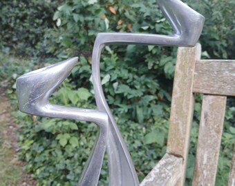 Puede incluir: Una escultura abstracta plateada con un diseño curvo y fluido. La escultura tiene puntas angulares y un acabado metálico liso. Está colocada al aire libre, sobre un banco de madera, con un fondo de follaje verde.