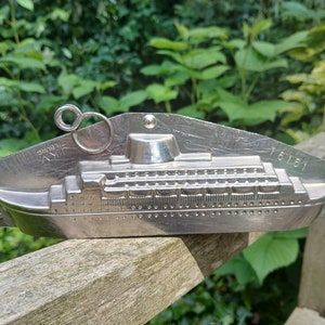 May include: A silver metal mould for making chocolate in the shape of a cruise ship. The mould has a loop handle and the text "16161" and "L.M.V.  Tilburg" is engraved on the side.