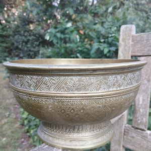 Puede incluir: Un cuenco grande de latón antiguo con intrincados patrones grabados. El cuenco presenta un borde ancho y una base de pedestal. Los diseños grabados incluyen formas geométricas y motivos florales. El cuenco se exhibe al aire libre sobre una superficie de madera.