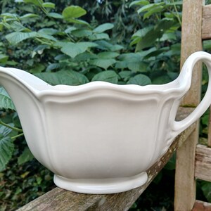 Può includere: Salsiera in ceramica bianca con bordo ondulato e un'unica maniglia. La salsiera è appoggiata su una superficie di legno.