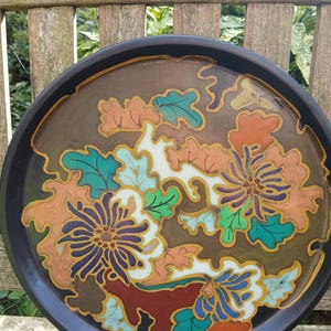 May include: A round, black decorative plate with a floral design. The flowers are in shades of orange, blue, and purple, with green leaves and gold accents. The plate has a brown center and a black rim.