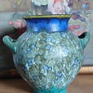 May include: A ceramic vase with two side handles, featuring a wide mouth and a rounded body. The glaze displays a vibrant blue rim transitioning into a mottled green and blue textured pattern across the surface, ending in a solid teal base.