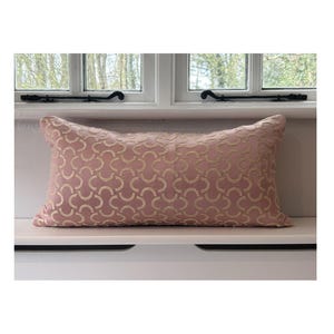 May include: A rectangular decorative pillow in a soft pink hue, featuring a repeating gold-colored geometric pattern. The pillow is placed on a white surface, with a window in the background.