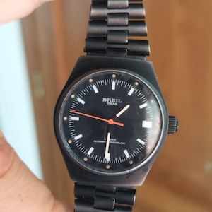 Puede incluir: Reloj de pulsera Breil Okay negro con una correa de metal negro. La esfera del reloj es negra con marcadores y agujas de hora blancos, y un segundero naranja. El reloj tiene una ventana de fecha y una corona en el lado derecho.