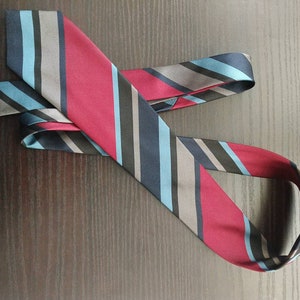 Puede incluir: Una corbata a rayas con rayas rojas, azules, grises y negras. La corbata está doblada por la mitad y colocada sobre una superficie de madera.