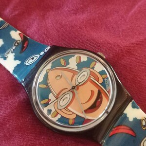 Może przedstawiać: Czarno-biały zegarek Swatch z kreskówkową twarzą na tarczy. Twarz jest żółta, pomarańczowa i niebieska na białym tle. Pasek zegarka jest niebieski, biały i czerwony z kreskówkowym wzorem.