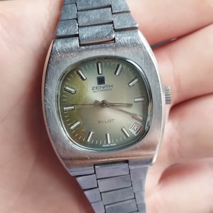 May include: A silver Zenith Pilot automatic wristwatch with a square face and a silver metal band. The watch face is a light green color with black markings and the words "Zenith Automatic" and "Pilot" printed on it. The date window is at the 3 o'clock position and shows the number 13.