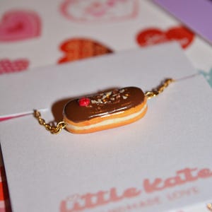 Könnte beinhalten: Eine goldene Kettenkette mit einem Miniatur-Schokoladen-Éclair-Anhänger. Der Éclair ist mit einem roten Bonbon und Streuseln verziert. Die Kette wird auf einem weißen Hintergrund mit rosa Herzen präsentiert.