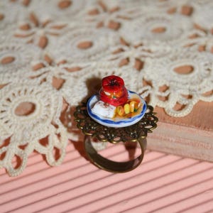 Puede incluir: Anillo en miniatura con un plato pequeño con comida roja, un cuadrado blanco y rodajas amarillas. El anillo tiene un engaste de filigrana de color bronce y una banda sencilla. El fondo es una superficie rosa con un tapete de encaje.