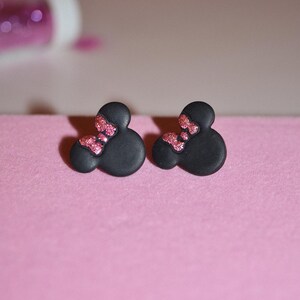 Puede incluir: Un par de pendientes de botón negros con detalles de purpurina rosa en forma de cabeza de Mickey Mouse.