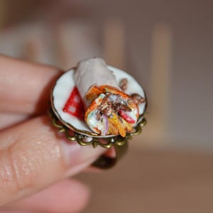 Puede incluir: Anillo único con una réplica detallada en miniatura de un gyro. El gyro está sobre un plato blanco pequeño con un patrón de cuadros rojos y blancos. El anillo tiene un acabado en bronce antiguo.