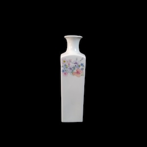 Puede incluir: Un jarrón blanco rectangular con la parte superior ensanchada. El jarrón presenta un diseño floral en colores pastel, incluyendo rosa, azul y morado. El jarrón es probablemente de cerámica o porcelana y es adecuado para exhibir flores.