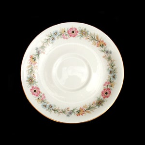 Pode incluir: Um pires de cerâmica branca com um design floral. O pires apresenta um anel de flores rosa, azuis, amarelas e verdes ao redor da borda. Uma borda dourada acentua a borda. Em um fundo preto.