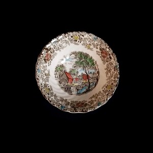 May include: A white ceramic bowl with a detailed landscape scene in the centre, featuring a red building, trees, and a waterfall. The rim has a gold trim and floral patterns in various colours.