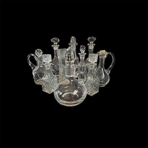 May include: An assortment of clear glass decanters and bottles, each with a unique shape and stopper. Some have handles, while others display diamond-cut patterns. The items are arranged against a black backdrop.
