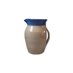 Op de afbeelding: Een keramische kan met een beige body en een blauwe rand. De kan heeft een gebogen handvat en een tuit om uit te schenken. Het ontwerp is eenvoudig en klassiek, geschikt voor het serveren van drankjes of als decoratief stuk.