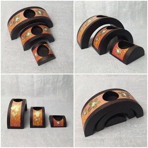 K&ouml;nnte beinhalten: Set aus drei unterschiedlich gro&szlig;en Kerzenhaltern aus Holz. Die dunkelbraunen Halter haben ein geschwungenes Design mit einem rosa Band mit goldenen und wei&szlig;en Blumenmustern. Jeder hat eine kreisf&ouml;rmige &Ouml;ffnung f&uuml;r Kerzen.