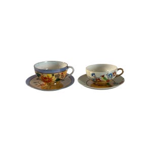 May include: Two vintage teacups and saucers. One cup has a blue rim and floral design with orange, yellow, and green hues. The other cup has a gold handle and a floral pattern with blue, red, and yellow colours. Both are on matching saucers.