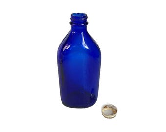 May include: A cobalt blue glass bottle with a rounded body and a narrow neck. A silver and white bottle cap sits next to the bottle. The bottle is empty and has a vintage aesthetic.