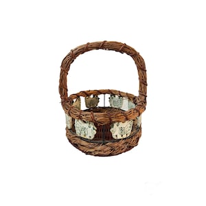 May include: A woven brown wicker basket with a handle. The basket features decorative white ceramic pieces with green designs around the rim. The handle is also made of woven brown wicker.