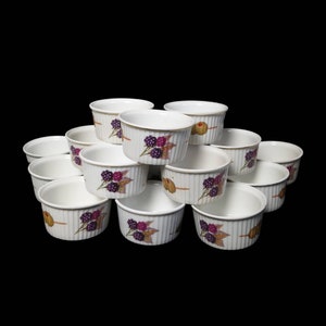 Può includere: Una collezione di ramequin in ceramica bianca con bordo dorato e design a coste verticali. Ogni ramequin presenta un disegno dipinto di more e olive. I ramequin sono impilati su uno sfondo nero.