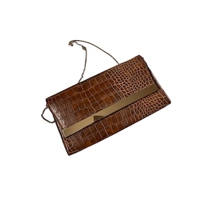 Könnte beinhalten: Eine braune Clutch mit Krokodilleder-Textur und einem goldfarbenen Metallstab auf der Vorderseite. Die Clutch hat einen silbernen Kettenriemen. Die Tasche ist rechteckig und scheint aus Leder zu sein.