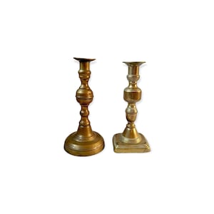 Puede incluir: Dos candelabros antiguos de latón. Uno tiene una base redonda y un centro bulboso, mientras que el otro tiene una base cuadrada. Ambos tienen un diseño clásico y ornamentado y están hechos de metal de color dorado cálido.