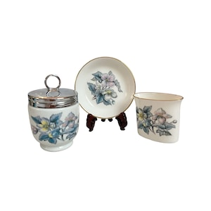 May include: A white porcelain set featuring a lidded jar with a silver-tone metal lid and ring handle, a small round dish, and an oval-shaped container. Each piece is decorated with a floral pattern of blue and pink flowers with green leaves.