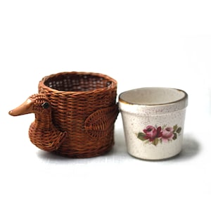 May include: A woven brown duck-shaped basket and a small white ceramic pot with a floral design. The duck basket has a detailed beak and eye. The pot features a gold rim and a cluster of pink roses.