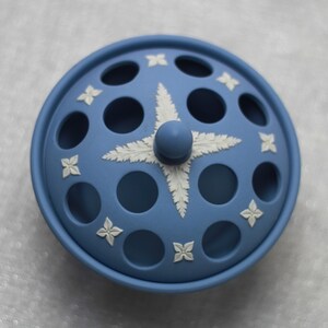 May include: A light blue ceramic container with a lid featuring a central starburst design and floral accents. The lid has circular openings and a small knob. The container is a decorative piece.